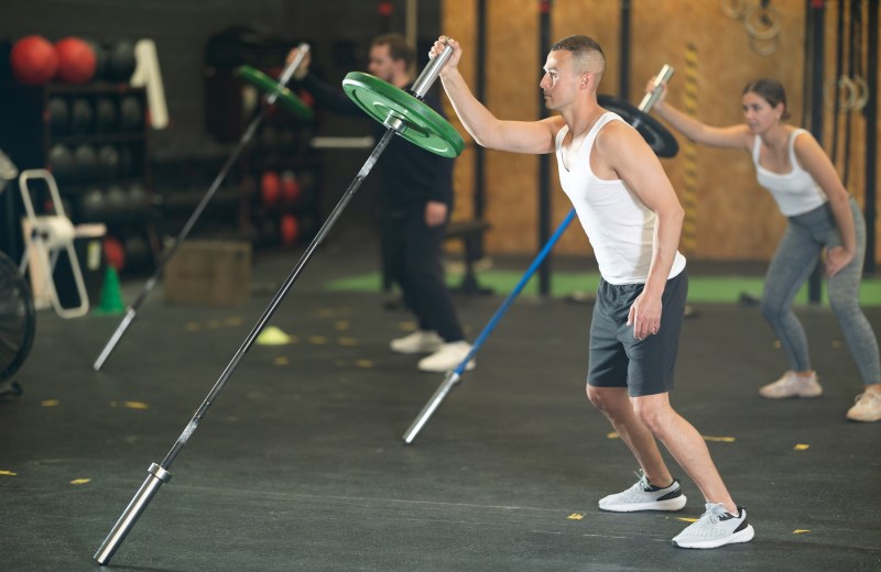 Man and people in gym doing standing one-arm landmine press exercise with barbell landmine attachment