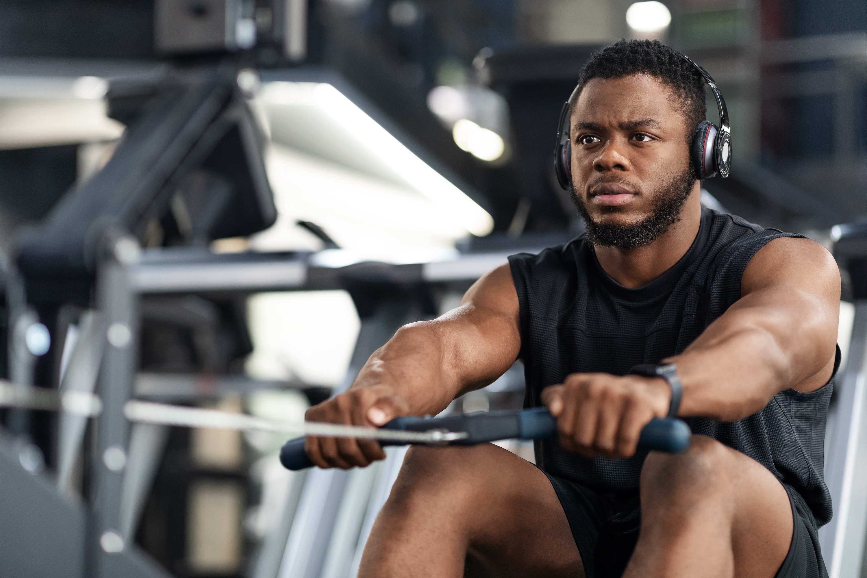 Closeup of handsome african american sportsman training body on modern rowing exerciser in gym, copy space. Motivated young black sportsman with wireless headset having workout on rowing machine