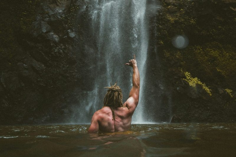 Shirtless man fist in the air punching air by a waterfall outside in nature back muscles